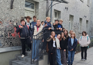 Uczniowie klasy VI pozują do zdjęcia na Pasażu Róży, na tle budynku wyłożonego szkłem. Wszyscy stoją i uśmiechają się do zdjęcia.
