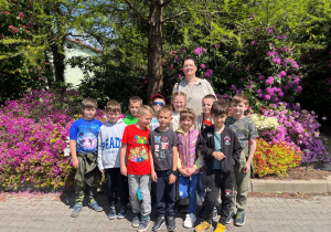 Uczniowie klasy pierwszej podczas wycieczki pozują do zdjęcia wraz z wychowawczynią na tle pięknych różowych rododendronów. Wszyscy uczniowie i wychowawca mają radosne miny podczas słonecznego i ciepłego dnia.
