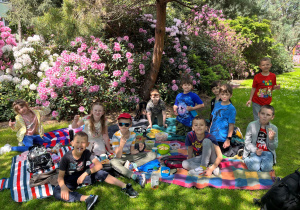 Radośni uczniowie klasy pierwszej siedzą na kocach w ogrodzie w gospodarstwie szkółkarskim. Jedzą przepyszne babeczki, ciasta, owoce, piją napoje. Wokół dzieci jest przepiękny krajobraz białe i różowe rododendrony , zielona trawa i kwitnące stokrotnki.