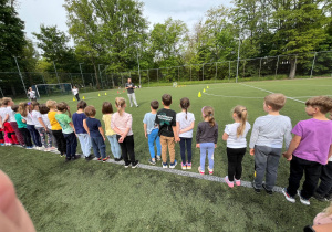 Przywitanie wszystkich uczestników imprezy. Przedszkolaki stoją w rzędzie na wprost nauczyciela prowadzącego zajęcia na boisku. Słuchają zasad.