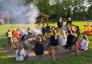 Uczniowie zgromadzeni wokół ogniska. Pieką kiełbaski, rozmawiają, śpiewają. Są radośni.