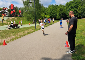 Na zdjęciu nauczyciel wychowania fizycznego i uczniowie biegnący na linię mety w pobliskim parku.