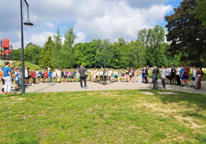 Uczniowie naszej szkoły na dużym placu w parku słuchają zasad przedstawianych przez nauczyciela wychowania fizycznego.