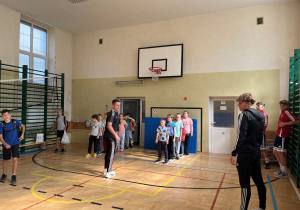 Trener wraz z podopiecznymi oraz zawodnik ŁKS witają się znaszymi uczniami. Dzieci ustawione w rzędach do rozgrzewki, słuchają uważnie jakie ćwiczenia będą wykonywać