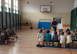 Uczniowie klas I-III na spotkaniu z panią policjantką w sali gimnastycznej. Dzieci z zaciekawieniem słuchają o bezpieczeństwie w ruchu drogowym