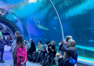 Uczniowie naszej szkoły podczas zajęć w Orientarium, słuchający z zaciekawieniem pracownika. Uczniowie fotografują rózne gatunki ryb.