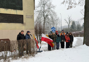 W zimowej scenerii na tle budynków stoi grupa uczniów z tabliczką SP40, obok są biało - czerwone flagi podkreślające patriotyczny charakter uroczystości