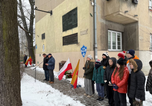 W zimowej scenerii na tle budynków stoi grupa uczniów z tabliczką SP40, za nimi są biało- czerwone flagi,scena oddaje atmosferę zadumy, pamięci i szacunku do historii