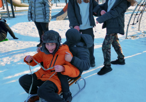 W tle zdjęcia plac zabaw. Na sankach siedzi dwóch uczniów - jeden z uczniów jest odwrócony od obiektywu natomiast drugi patrzy w obiektyw i jest uśmiechnięty. Nad nimi stoją trzy dziewczyny - jedna w oddali, a dwie bezpośrednio przy chłopcach, które śmieją się.