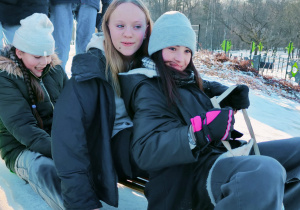 Na sankach siedzą dwie dziewczyny nie patrzą w obiektyw , ale się uśmiechają. Za sankami siedzi jedna uśmiechnięta dziewczyna i nad nią stoi jeszcze jedna dziewczyna, która próbuje popchnąć sanki z górki. Wszystko jest na tle zimowej scenerii parku.