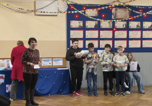 Grupa nagrodzonych chłopców stoi w rzędzie z upominkami, którzy zostali nagodzeni za konkurs organizowany z okazji Dnia Babci i Dziadka. Pani Sylwia organizatorka konkursu gratuluje laureatom.