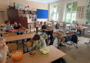 Uczniowie w sali lekcyjnej siedzą przy swoich ławkach, a przed sobą zamiast książek mają opakowania bądź miski z popcornem
