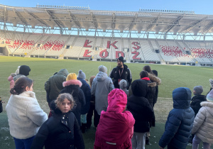 Stadion ŁKS, pan opowiada o murawie, jak o nią dbają, i że nie można wchodzić na nią w obuwiu jakim się chce. Dzieci odwrócone tyłem do aparatu, słuchają pana