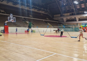 Dzieci na hali sportowej MOSiR, tu rozstawione są siatki do grania w badmintona, dziewczynka biega wokoło, po lewej stronie chłopiec rzuca do celu