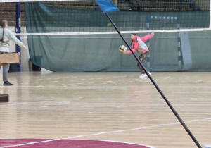 Dzieci na hali sportowej MOSiR, dziewczynka w jednym ręku ma piłkę a drugą ręką chce zaserwować, po drugiej stronie siatki jest druga dziewczynka przygotowana na odbicie piłki