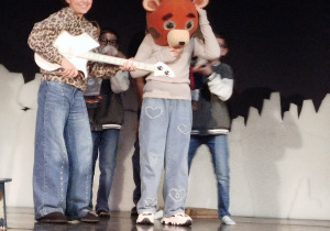 Scena teatru, a na niej czworo uczestników półkolonii. Dziewczynka trzyma gitarę, inna ma maskę misia na głowie a za nimi tańczy dwóch chłopców