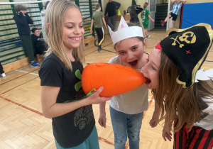 Sala gimnastyczna, uśmiechnięte dziewczynki udają, że nadgryzają ogromną, sztuczną marchewkę