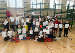 Sala gimnastyczna, wszyscy uczestnicy półkolonii pozują do zdjęcia z dyplomami półkolonisty, z uśmiechami na twarzach.