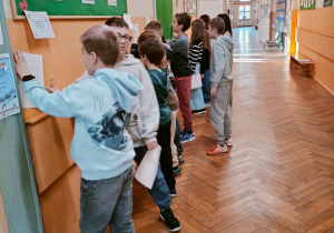 Uczniowie klasy 4 pracują na szkolnym korytarzu przy stanowiskach z zadaniami umieszczonymi na ścianie; dzieci czytają polecenia i zapisują odpowiedzi na kartkach.