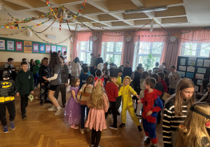 Uczniowie na korytarzu szkolnym podczas zabawy karnawałowej, są przebrani za różne postacie zadowoleni bawią się w rytm muzyki.