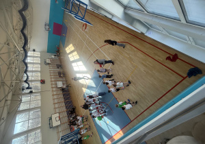 Zdjęcie sali gimnastycznej wykonane z góry, Zawodnicy stoją w rzędach przygotowują się do kolejnej konkurencji, rozmawiają ze swoimi trenerami.