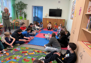 Na zdjęciu uczniowie w świetlicy siedzący w kręgu. Jedna z dziewczynek wyciąga rękę do kota. Po lewej stronie na zdjęciu pani Iza założycielka fundacji