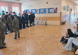 Na korytarzu drugiego piętra klasa VIII spotkała się z uczniami szkoły ponadpodstawowej ,,Oświatowiec". Uczniowie wspomnianej szkoły ubrani w wojskowe mundury stoją na baczność przed uczniami klasy VIII, którzy siedzą na krzesłach