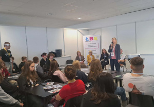 Uczniowie klas VII i VIII siedzą przy stolikach i Pani prowadząca wykład, przemawia do uczniów przez mikrofon, wokoło plakaty o przyszłości edukacyjnej.