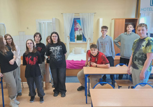 Uczniowie pozują do zdjęcia w specjalnie przygotowanej sali na styl hotelowej .W tle okno, łózko hotelowe, łazienka z prysznicem oraz szafa