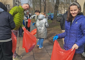 Uczennice i pani od angielskiego trzymają czerwone worki i sprzątają teren Osiedla Montwiłła - Mireckiego. W tle bloki i roślinność.