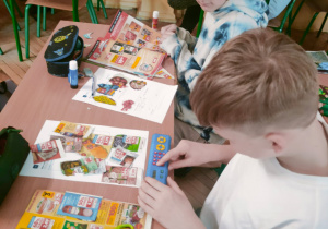 Dwóch uczniów przy ławce skrupulatnie planuje swoje zakupy, korzystając z gazetek reklamowych