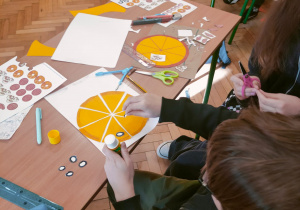 Uczniowie klasy 4 przy szkolnej ławce wycinają i przyklejają papierowe składniki oraz matematyczne symbole, tworząc „matematyczną pizzę”.