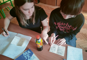 Uczennice klasy 8 przy szkolnej ławce rozwiązują skomplikowane zagadki logiczne i matematyczne szyfry
