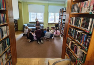 Uczniowie naszej szkoły siedzą na wykładzinie w bibliotece wokół pani prowadzącej zajęcia i pani czyta uczniom fragment tekstu