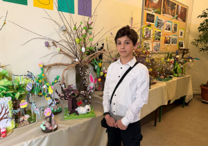 Uczeń klasy V stoi przy stole nakrytym białym obrusem obok niego stoi praca, którą wykonał. Wokoło jego pracy prezentowane są jeszcze inne prace uczniów.