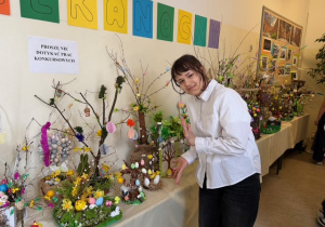 Uczennica klasy V stoi przy stole nakrytym białym obrusem obok niej stoi praca, którą wykonała. Wokoło jej pracy prezentowane są jeszcze inne prace uczniów.