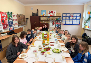 Uczniowie klasy V pozują do zdjęcia przy nakrytym stole. W tle wiszą tablice z wydarzeniami historycznymi. Świąteczne śniadanie - to jajka, kabanosy, soki .