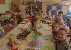 Uczniowie klasy pierwszej zgromadzeni przy pięknie nakrytym stole z licznymi smakołykami pozują do zdjęcia z uśmiechami na twarzy. Stół przygotowany w sali lekcyjnej klasy 1.