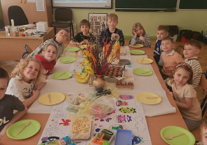 Uczniowie klasy pierwszej z drugiej strony zgromadzeni przy pięknie nakrytym stole z licznymi smakołykami pozują do zdjęcia z uśmiechami na twarzy. Stół przygotowany w sali lekcyjnej klasy 1.