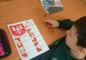 Uczeń w szkolnej ławce koloruje czerwoną kredką kartę pracy z matematyki. Pokolorowane kratki tworzą zarys polskiego godła – orła w koronie. Obok leży podręcznik "Matematyka 5".