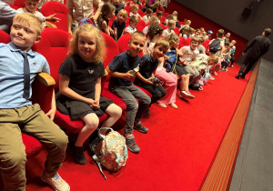 Uczniowie klasy I w teatrze siedzą na krzesełkach tuż przed sceną. Uczniowie z uśmiechami pozują do zdjęcia, wszyscy elegancko ubrani, grzecznie siedzą na czerwonych krzesełkach.