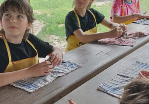 Uczniowie siedzą przy stole pod wiatą, Przed nimi leżą ściereczki a uczniowie ściskają w rękach glinę aby ją uformować.