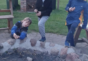 Uczniowie podczas ogniska. Dwóch stoi przy ognisku i patrzy w prawą stronę, jeden kuca przy ognisku dorzucając patyk