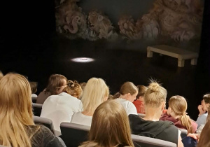 Publiczność w siedzi fotelach teatralnych, wszyscy skupieni na scenie. Scena przygotowana pod przedstawienie, są na niej rekwizyty