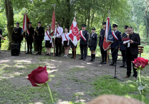 Park im. Józefa Piłsudskiego, uczniowie klasy siódmej stoją ze sztandarem szkoły przy kamieniach upamiętniających wydarzenia sprzed lat. Obok uczniów przemawia urzędnik państwowy, uczniowie stoją między służbami mundurowymi