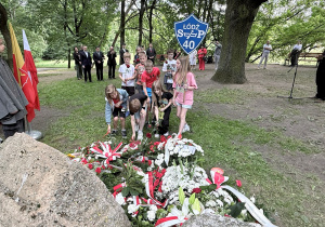 Uczniowie klasy czwartej z szyldem szkoły stawiają znicz przed kamieniami upamiętniającymi rewolucjonistów w parku i. J. Piłsudskiego. W tle służby mundurowe oraz harcerze i osoby reprezentujące miasto Łódź.