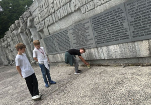 Uczniowie pod pomnikiem Rewolucjonistów w Parku im. Józefa Piłsudskiego. Dwóch uczniów stoi, trzeci stawia znicz pod pomnikiem.