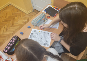 Dwie uczennice siedzą przy szkolnej ławce i rozwiązują zadania podczas matematycznej gry planszowej. Jedna z nich trzyma długopis nad planszą z zadaniami, a druga obserwuje grę. Na ławce znajdują się również zeszyty, piórniki i przybory szkolne.