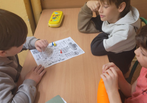 Troje uczniów siedzi przy szkolnej ławce i uczestniczy w matematycznej grze planszowej. Na stole leży papierowa plansza z polami zawierającymi zadania matematyczne oraz kolorowymi pionkami. Jeden z uczniów przesuwa rękę nad planszą, a pozostali obserwują przebieg gry.